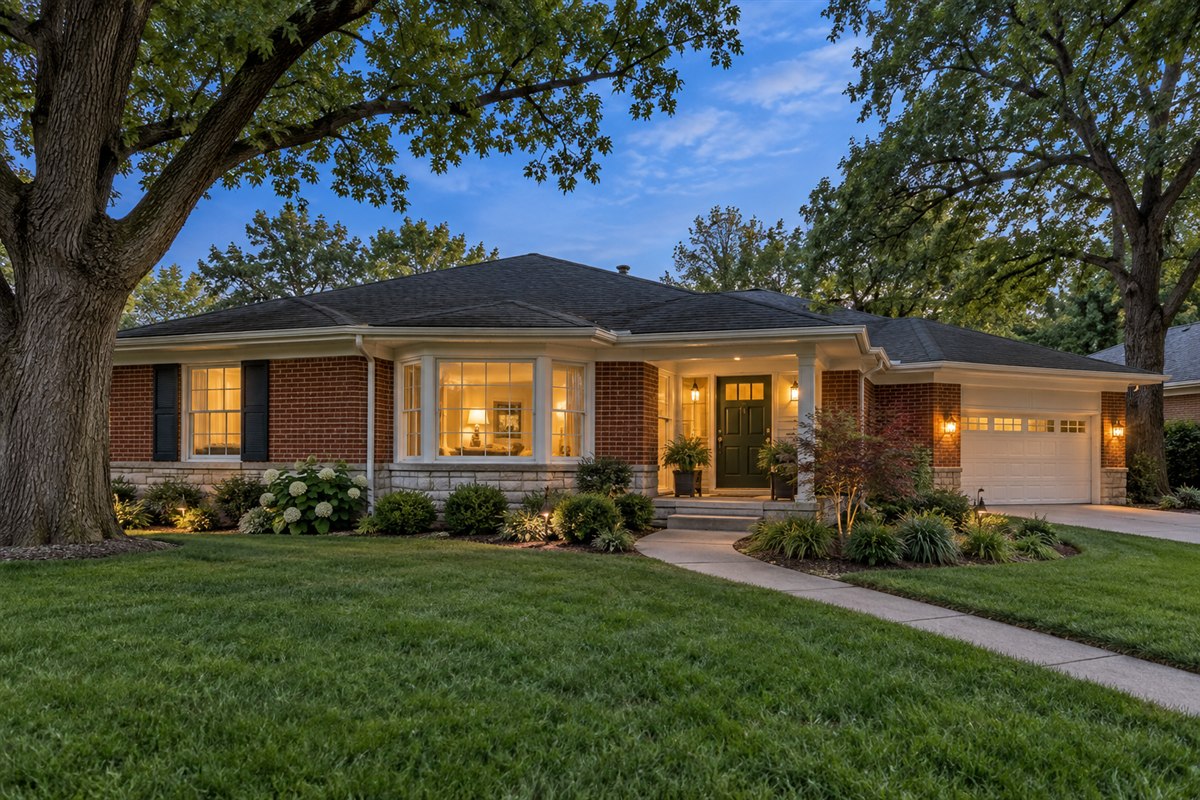 Red-brick ranch home with white trim, a front bay window and mature trees at dusk